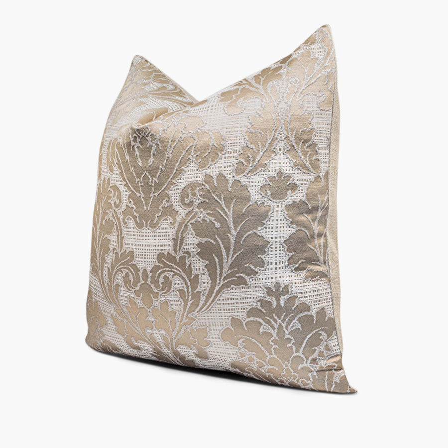 A champagne-beige damask throw pillow sits against a white background. The damask pattern is outlined in metallic-silver thread and set against a white, woven criss-cross base. The pillow is shown from the side.