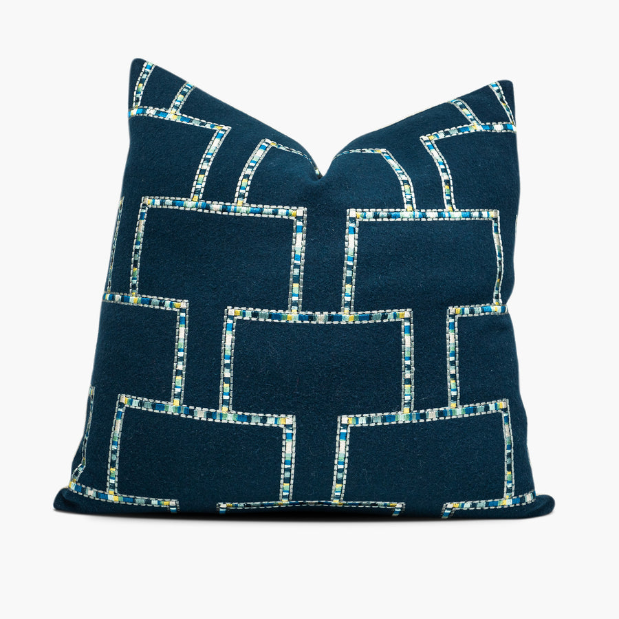 A dark blue, wool throw pillow is shown from the front. The pillow has an embroidered geometric motif in shades of blue, silver, and yellow.