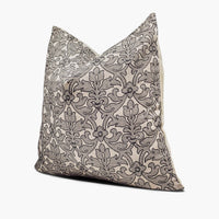 A tan throw pillow sits against a white background. The pillow has modern damask pattern embroidered in black. The pillow is shown from the side.