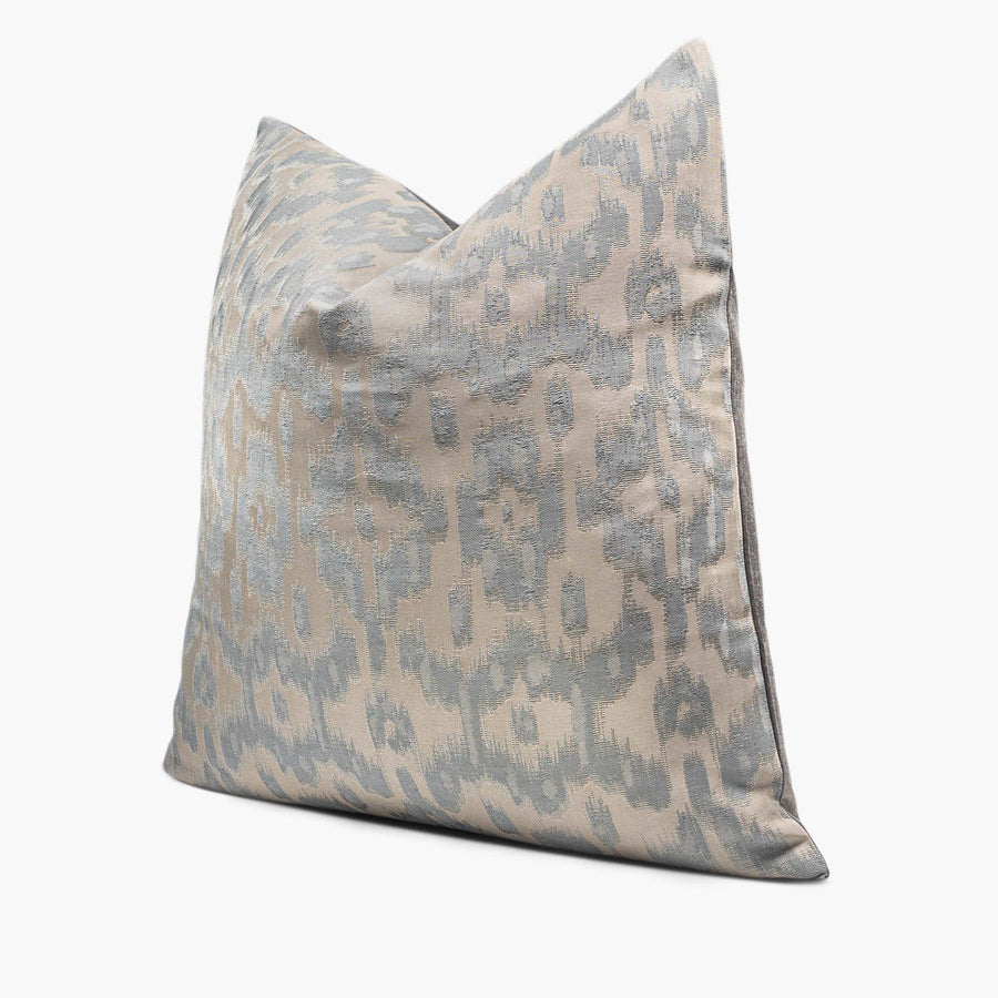 A throw pillow with a silvery ikat motif sits against a white background. The pillow is shown from the side.