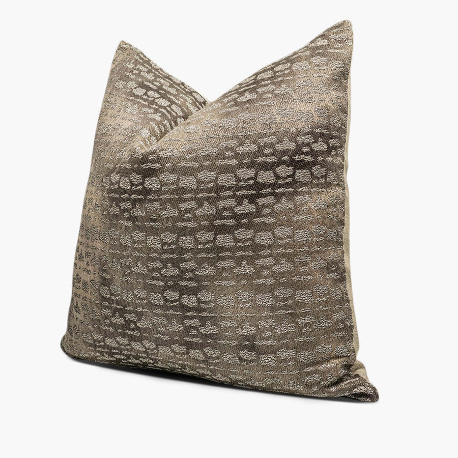 A taupe-brown throw pillow sits against a white background. The pillow has an abstract, spotted motif. The spots are metallic silver. The pillow is shown from the side.