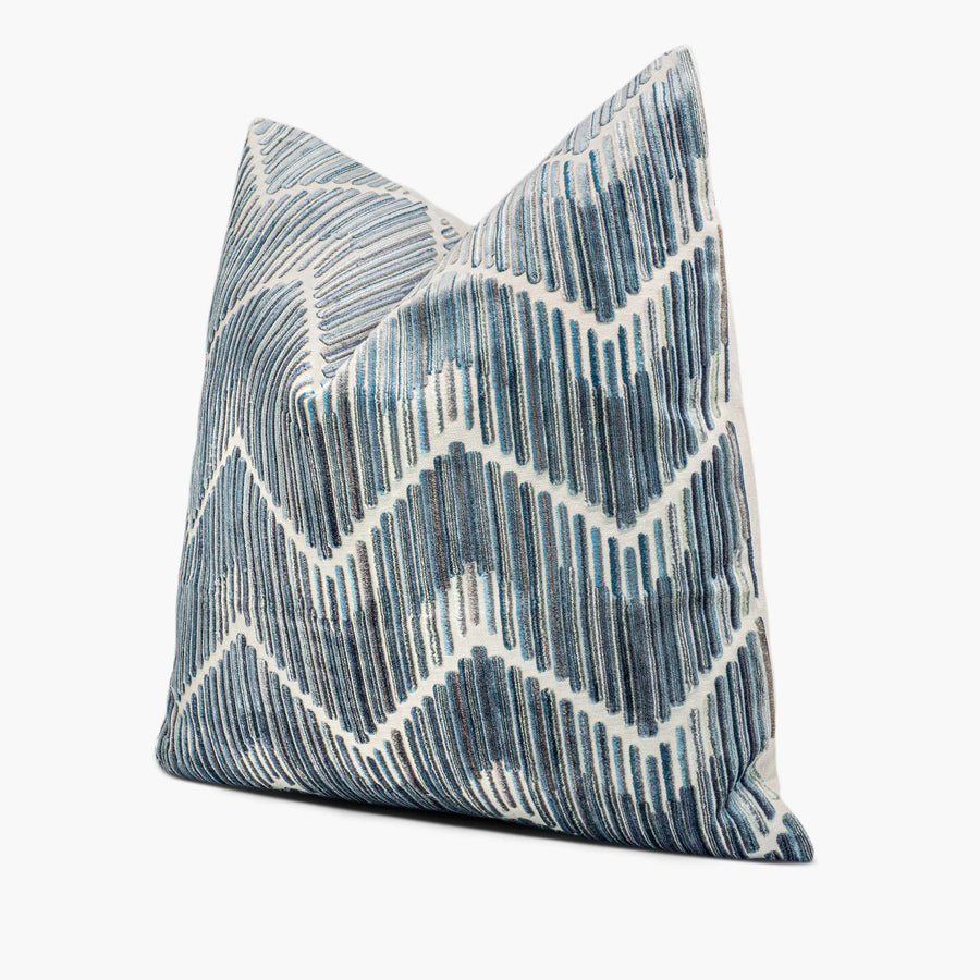 A velvet throw pillow sits against a white background. The throw pillow has a flame stitch chevron motif in shades of steel blue. The pillow is shown from the side.