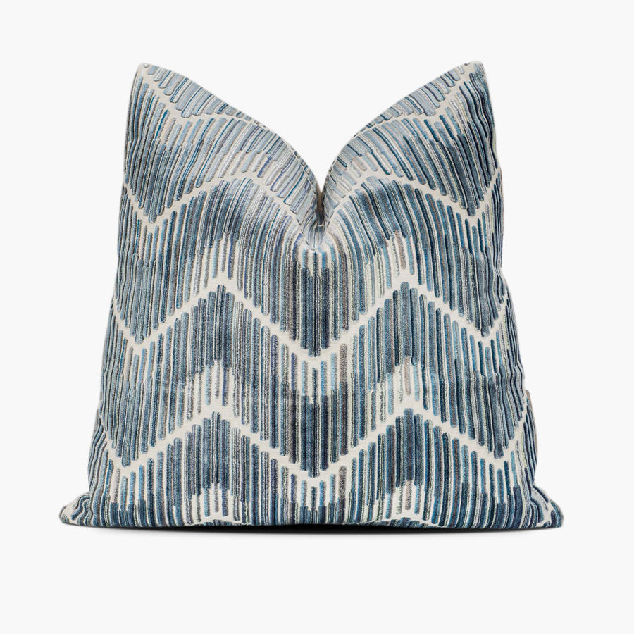 A velvet throw pillow sits against a white background. The throw pillow has a flame stitch chevron motif in shades of steel blue.